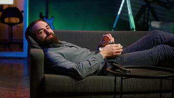 Smiling man lounged on couch eating popcorn while watching movie on streaming service, enjoying time off. Upbeat person laying down in apartment consuming snacks, looking at films, camera A video