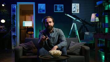 Man relaxing at home on couch, stuffing mouth with popcorn while watching movies on TV in apartment. Person eating snacks and streaming TV shows in neon lit living room, camera B video