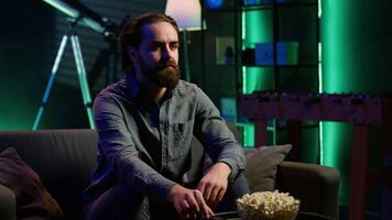 Man relaxing at home on couch, stuffing mouth with popcorn while watching movies on TV in apartment. Person eating snacks and streaming TV shows in neon lit living room, camera A video