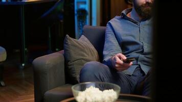 Smiling person on couch picking movie on streaming service using remote and texting on mobile phone. Happy man watching shows on television, zapping though channels, chatting smartphone, camera A video