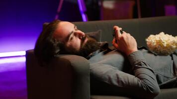 Gamer lounged on couch engaging in multiplayer game using gamepad, eating snack. Person in home theatre playing videogames on gaming console with bowl of popcorn in lap, camera A video