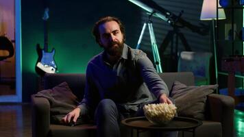 Man turning on TV using remote control, enjoying leisure time at home, munching on popcorn. Couch potato person in neon illuminated apartment eating salty snack, entertained by television, camera A video