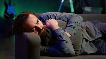 Man laying down hugging pillow watching TV, heartbroken by upsetting movie plot. Sad viewer lounged on couch in RGB illuminated apartment, upset by depressing television show scene, camera A video