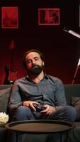Vertical Bored gamer gaming on TV console using gamepad, eating popcorn, living sedentary lifestyle. Lazy man in home theatre playing videogames and consuming snacks, sitting on couch, camera B video
