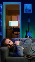 Vertical Man laying down hugging pillow watching TV, heartbroken by upsetting movie plot. Sad viewer lounged on couch in RGB illuminated apartment, upset by depressing television show scene, camera B video
