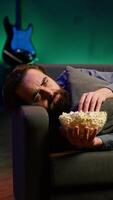 Vertical Depressed man at home lounged on couch feeling low, seeking comfort by watching movies on TV and eating popcorn. Glum person laying down, suffering from depression, watching TV to feel better, camera A video