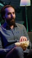 Vertical Amused person sitting on couch using TV to binge comedy series on subscription based streaming services, enjoying popcorn. Man laughing at funny shows on television, eating snack, camera A video