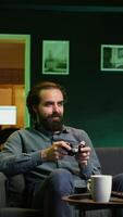 Vertical Joyful gamer at home gaming and enjoying leisure time. Smiling man sitting on couch in neon lit living room, excited to play videogame on console, relaxing after hard day at work, camera B video