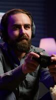 Vertical Excited man playing videogames using controller, having fun in neon illuminated room. Enthusiastic gamer competing in multiplayer game using gamepad, talking with friends through headset, camera A video