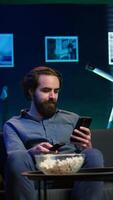 Vertical Smiling person on couch picking movie on streaming service using remote and texting on mobile phone. Happy man watching shows on television, zapping though channels, chatting smartphone, camera B video