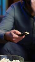 Vertical Close up of man in dimly lit apartment using remote control to browse though movies on streaming service. Remote control used by person in living room flipping through channels on TV, camera A video