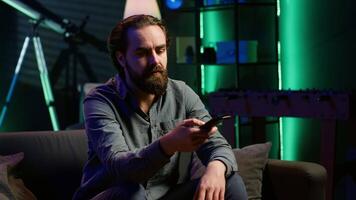 Bored person on couch in front of TV binging series on subscription based streaming services, browsing using remote. Weary man watching VOD shows on television set, zapping though channels, camera A video