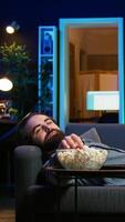 Vertical Smiling man lounged on couch at home eating popcorn while browsing though movies available on streaming service, enjoying time off from work. Man laying down in apartment consuming snacks, camera B video