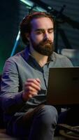 Vertical Man typing on laptop keyboard in neon lit apartment, sitting on couch and watching TV with bowl of popcorn ready. Teleworking person solving tasks on notebook with film playing in background, camera A video
