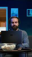 Vertical Man watching TV in apartment office and solving business related tasks on laptop, working from home. Freelancer on couch in living room looking at movies and working on notebook device, camera B video