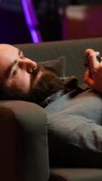 Vertical Gamer lounged on couch engaging in multiplayer game using gamepad, eating snack. Person in home theatre playing videogames on gaming console with bowl of popcorn in lap, camera A video