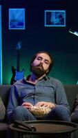 Vertical Fatigued man watching shows on TV late at night and munching on popcorn, paying close attention to screen. Tired sedentary man sitting in pajamas all day consuming mindless entertainment, camera B video