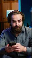 Vertical Man sitting on couch at home texting friend on smartphone, inviting him to watch movie on streaming service. Person at home discussing with mate on mobile phone, recommending TV show, camera A video