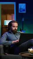 Vertical Teleworker writing emails on laptop at home while watching TV. Remote employee working from home in cozy apartment personal office, typing on notebook, turning on TV using remote control, camera B video