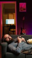 Vertical Gamer lounged on couch engaging in multiplayer game using gamepad, eating snack. Person in home theatre playing videogames on gaming console with bowl of popcorn in lap, camera B video