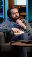 Vertical Sad man on couch eating popcorn from stress while watching heartbreaking drama movie on streaming service. Person consuming snack, distressed by TV show favorite character dies, camera A video