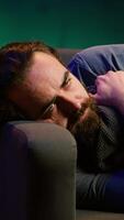 Vertical Man laying down hugging pillow watching TV, heartbroken by upsetting movie plot. Sad viewer lounged on couch in RGB illuminated apartment, upset by depressing television show scene, camera A video