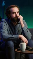Vertical Man at home shocked by thriller film on TV, drinking coffee. Stupefied person at home left with eyes wide open by dramatic plot twist, watching suspenseful movie on TV, enjoying beverage, camera A video