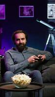 Vertical Cheerful gamer sitting on couch engaging in online competitive multiplayer tourney using gaming console and gamepad. Upbeat player engaging in fun videogames, enjoying leisure time, camera B video