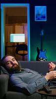 Vertical Smiling man lounged on couch eating popcorn while watching movie on streaming service, enjoying time off. Upbeat person laying down in apartment consuming snacks, looking at films, camera B video