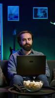 Vertical Man typing on laptop keyboard in neon lit apartment, sitting on couch and watching TV with bowl of popcorn ready. Teleworking person solving tasks on notebook with film playing in background, camera B video