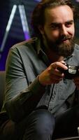 Vertical Excited man holding controller in neon illuminated apartment, playing videogames. Happy gamer on couch using joystick to participate in PvP game on console system, relaxing and having fun, camera A video