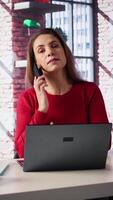 Vertical Female freelancer at home calling her team members to discuss online research and surfing the internet, utilizing a laptop for telework. Staying connected on telephone line call. Camera B. video