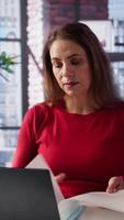 Vertical Focused mature woman multitasking on a laptop in her stylish loft, connected to the internet while managing freelance work and remote responsibilities. Browsing web at home office. Camera B. video