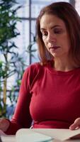 Vertical Focused mature woman multitasking on a laptop in her stylish loft, connected to the internet while managing freelance work and remote responsibilities. Browsing web at home office. Camera A. video