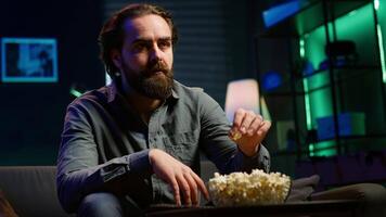 Puzzled man eating popcorn while watching confusing movie on streaming service, enjoying time off from work. Person consuming snacks, looking at film broadcasted on TV with convoluted plot, camera A video