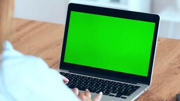 A person is focused on their laptop, which displays a plain green screen. The setting is a cozy workspace with a wooden desk and minimal clutter, creating a productive atmosphere. video