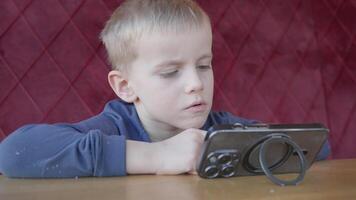 A 4-5-year-old European boy sits at the table, watching cartoons on his phone, leaning in closely. Immersed in digital entertainment, he focuses on the screen, enjoying his favorite animated content. video