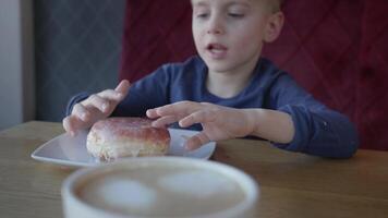 A 4-5 year old boy sits at the table, happily biting into a large, sweet donut. The boy is wearing a casual outfit, and the atmosphere is cozy and warm, perfect for a fun snack time. video