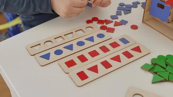 The child carefully arranges the red and blue wooden geometric shapes circle, square, triangle and rectangle on the table, sorting them by shape. A wonderful Montessori-inspired learning experience. video