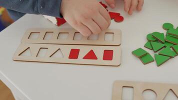 The child carefully arranges red wooden geometric shapes circle, square, triangle and rectangle on the table, sorting them by shape. A wonderful Montessori-inspired learning experience. video