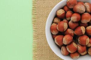 Hazelnuts in white bowl on green background. Bowl of hazelnuts with burlap border. Nuts in white bowl on green backdrop. Pile of hazelnuts in white container. Hazelnut in bowl with jute fabric photo