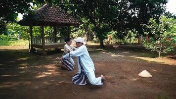 Two individuals performing a traditional dance in a lush outdoor setting, with a gazebo in the background. The dancers wear traditional attire and are engaged in a cultural performance video