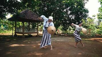 Two individuals in traditional attire performing a cultural dance or activity in a rural setting, with a gazebo and lush greenery in the background video