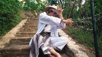 A person wearing a traditional conical hat and cultural attire performs a dance on stone steps surrounded by greenery. The dancer's hands are elegantly positioned, showcasing a graceful movement video