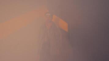 A young man stands against a wall with a yellow geometric design. He wears a colorful sweater and a shiny jacket, with goggles on his head. The scene has a misty atmosphere, adding a creative vibe video