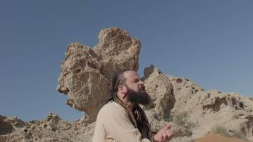 A man with a staff in his hand and the clothes of a shaman stands on a rock among the sands of the desert and dances video