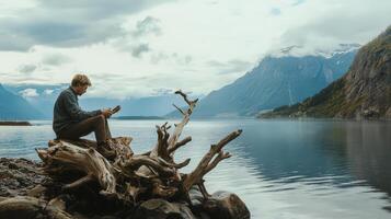 A man sitting on a log by a lake with mountains in the background photo