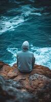 Person sitting on rocky shore watching ocean waves at dusk photo