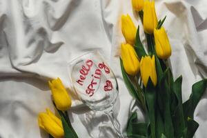 Hello SPRING message on wine glass with yellow fresh tulips on white bed with trendy deep shadows. Spring flowers in yellow colors. Romantic postcard wallpaper design Copy space photo