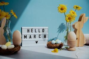 HELLO MARCH text on Lightbox with bunny wooden figures and fresh flowers. Easter composition content still life. Christian symbols of spring holiday photo